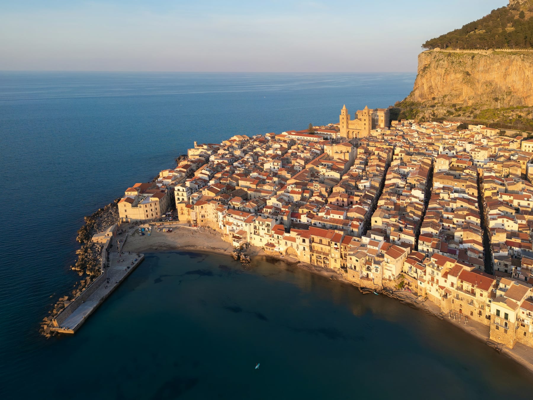 Aerial view of a historic Sicilian coastal town at golden hour, stone houses with terracotta roofs along the sea